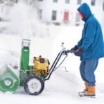 Person using a snow blower to clear snow from a residential driveway during winter.
