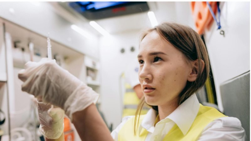 Healthcare trainee wearing a high-visibility vest and gloves carefully preparing a syringe inside an emergency medical vehicle.